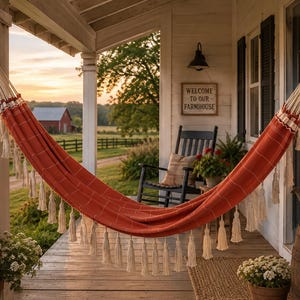 May include: A rust-coloured hammock featuring a grid pattern and cream-coloured fringe tassels along the bottom edge. The hammock is suspended between two white porch posts, with thick woven ropes at each end for secure attachment and support.