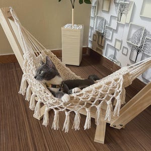 May include: A cream-colored macrame cat hammock suspended on a light-toned wooden frame. The hammock features a knotted rope design with decorative fringe along the edges. A grey and white cat with a patterned collar is resting inside the hammock.