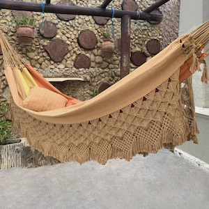May include: A beige fabric hammock featuring intricate macrame detailing along the edges with a diamond pattern and fringe. Inside the hammock, there is a solid orange pillow and a yellow fabric section, suspended by thick ropes from a wooden frame.