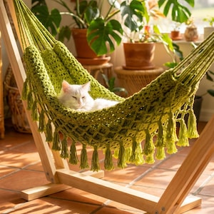 May include: A white cat resting in a lime green crocheted hammock suspended from a light-colored wooden stand. The hammock features a thick, open-weave pattern with decorative tassels hanging along the bottom edge, providing a cozy space.