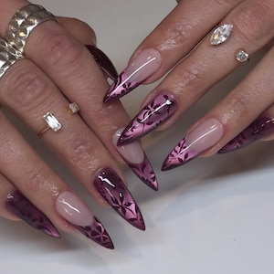 Puede incluir: Primer plano de uñas manicuradas con forma larga y puntiaguda. Las uñas presentan un diseño degradado, que transiciona de una base nude a un borgoña profundo con un patrón floral. Varios anillos adornan los dedos.