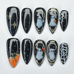 May include: A set of ten black press-on nails with Halloween-themed designs. The designs include gold stars, a spider on an orange pumpkin, a ghost, a jack-o'-lantern, and a white spiderweb. The nails have a glossy finish.