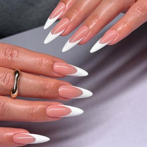 May include: Close-up of a hand with long, almond-shaped nails. The nails feature a classic French manicure design, with a pale pink base and crisp white tips. A gold ring is visible on one finger. The nails are well-manicured and glossy.