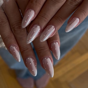 May include: Close-up of long, almond-shaped nails with a pearlescent finish. Each nail features small, evenly spaced white polka dots. The nails are a light, neutral colour, creating a delicate and stylish look.