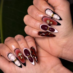 May include: Long, pointed acrylic nails with various designs. One nail features a white and pink marble design with gold accents. Another has a gradient of red to pink with a gold cross. Other nails are burgundy, tortoiseshell, and black and white.
