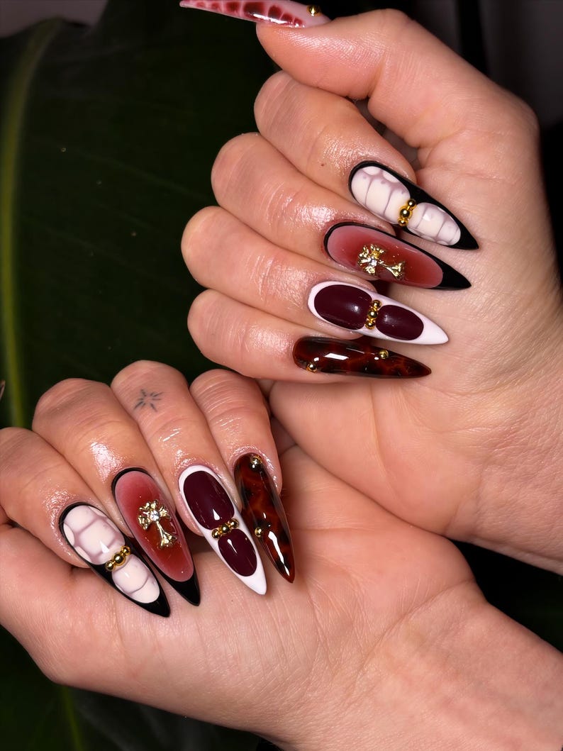 May include: Long, pointed acrylic nails with various designs. One nail features a white and pink marble design with gold accents. Another has a gradient of red to pink with a gold cross. Other nails are burgundy, tortoiseshell, and black and white.