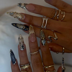 May include: Close-up of hands adorned with long, embellished nails and various silver and gold rings. The nails feature intricate designs with glitter, gems, and metallic accents. Rings include abstract shapes and delicate bands.