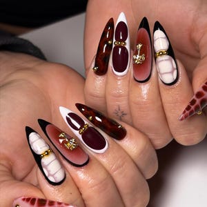 May include: A set of long, pointed fingernails with various designs. Some nails feature a dark red and brown marbled effect, while others have white tips, burgundy accents, and gold embellishments. One nail has a pink base with a red pattern.