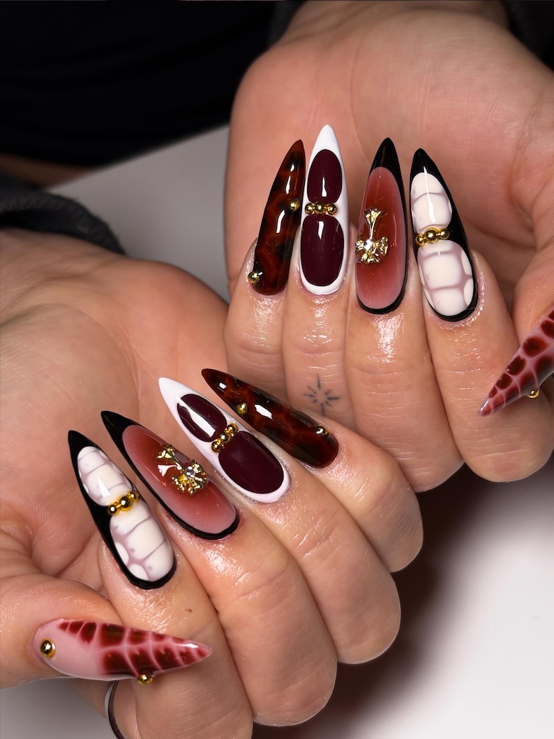 May include: A set of long, pointed fingernails with various designs. Some nails feature a dark red and brown marbled effect, while others have white tips, burgundy accents, and gold embellishments. One nail has a pink base with a red pattern.