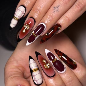 May include: A set of long, pointed acrylic nails. Designs include a white and pink pattern, a burgundy and white gradient with a gold cross, and a tortoiseshell pattern. Some nails have gold bead accents and black tips.