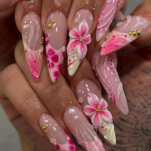 May include: Long, pointed acrylic nails with a pink and white floral design. The nails feature 3D flowers, gold accents, and a glossy finish. Some nails have a pink and white polka dot pattern.