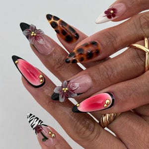 Puede incluir: Un conjunto de uñas artificiales largas en forma de almendra con varios diseños. Algunas uñas presentan un patrón de carey, mientras que otras tienen un diseño degradado rosa y rojo. Varias uñas están adornadas con adornos florales en 3D y tachuelas doradas. Las uñas tienen puntas negras.