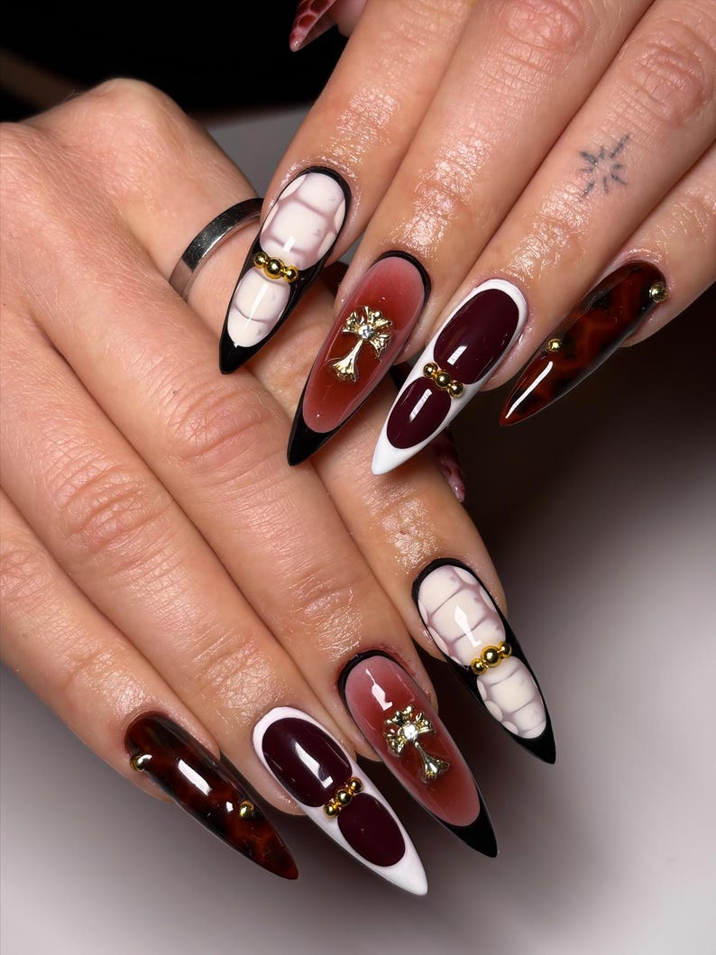 May include: Long, almond-shaped artificial nails with a variety of designs. Some nails feature a white base with a grid pattern and gold accents. Others have a gradient of red and white, with a gold cross or gold beads. The tips are black.