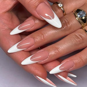 May include: Close-up of a hand with long, almond-shaped nails. Each nail features a classic French manicure design with a white tip and a nude base. Gold rings with gemstones adorn the fingers.