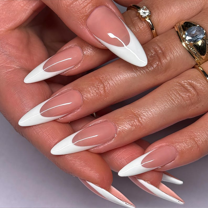 May include: Close-up of a hand with long, almond-shaped nails. Each nail features a classic French manicure design with a white tip and a nude base. Gold rings with gemstones adorn the fingers.
