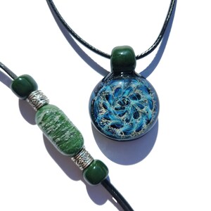May include: A round glass pendant featuring a swirling blue and white pattern on a black cord, accompanied by a bracelet strand with a textured green cylindrical bead, two smaller dark green spherical beads, and two silver-toned metal spacers.