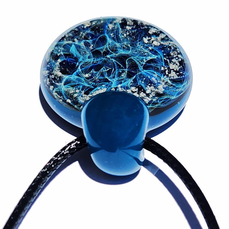 A round, blue urn pendant with a swirling, abstract design in shades of blue with mixed in ashes. The pendant is suspended from a black cord. The design is encased in clear glass, creating a unique, artistic piece.