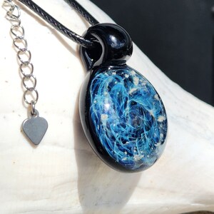 A black glass cremation pendant necklace with a blue and ashes swirling design. The pendant is oval-shaped and hangs from a black cord with a silver chain and heart-shaped charm. The pendant measures approximately 1.5 inches in length.
