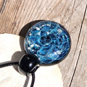 A round, cremation glass pendant with a swirling blue with ashes design, resembling a galaxy. The pendant is attached to a black cord and has a black, rounded clasp. The background is a light-colored, wooden surface.