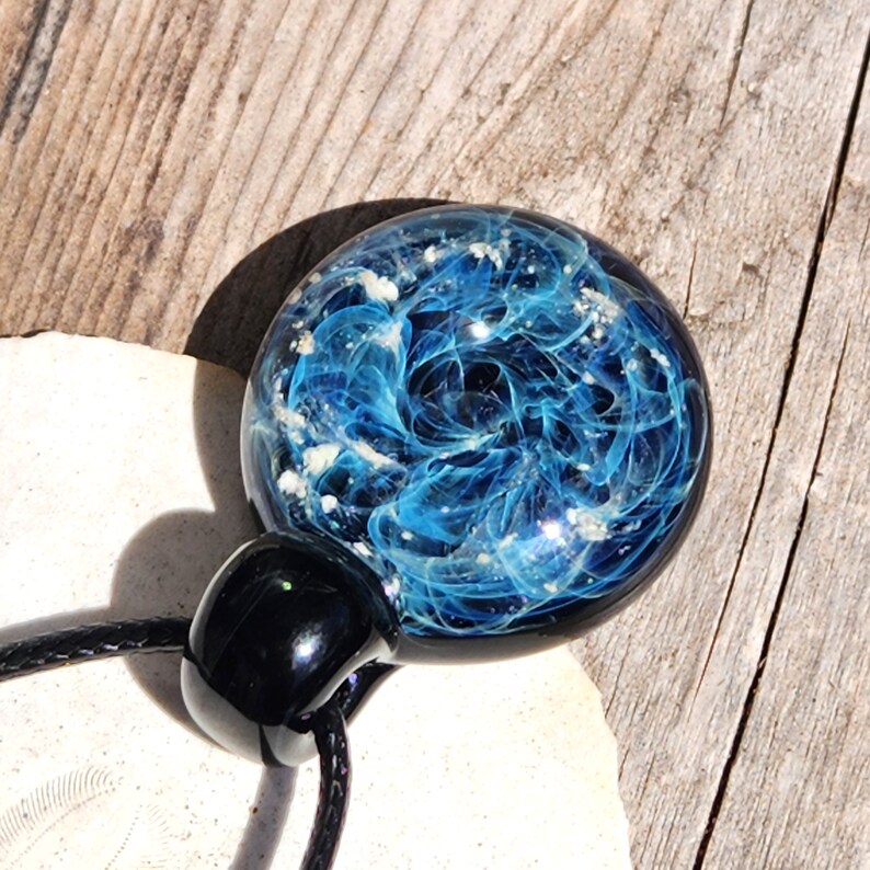 A round, cremation glass pendant with a swirling blue with ashes design, resembling a galaxy. The pendant is attached to a black cord and has a black, rounded clasp. The background is a light-colored, wooden surface.