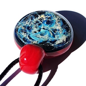 A handcrafted cremation glass pendant with a celestial design. The round pendant features swirling blue with infused ashes patterns encased in clear glass, set against a dark background. It has a red base and a red bail, strung on a black cord.