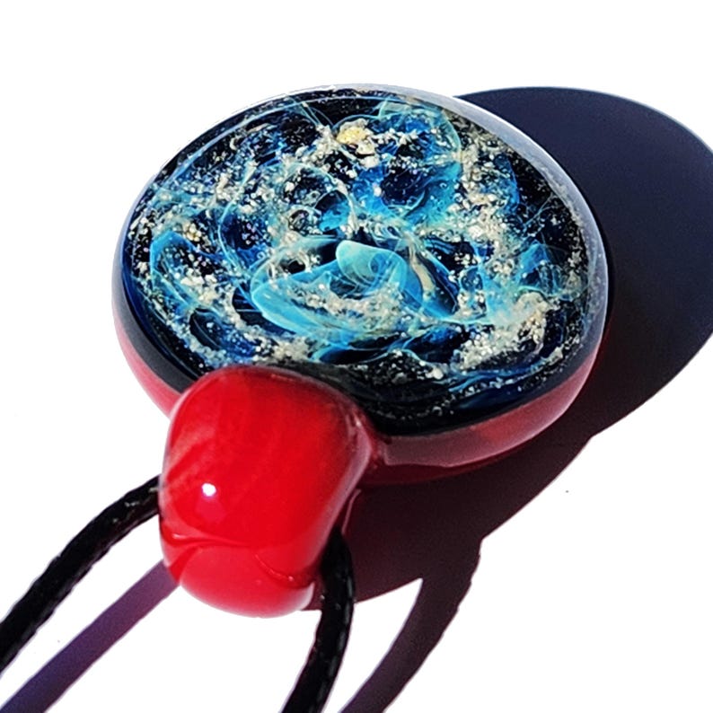 A handcrafted cremation glass pendant with a celestial design. The round pendant features swirling blue with infused ashes patterns encased in clear glass, set against a dark background. It has a red base and a red bail, strung on a black cord.