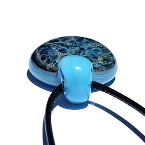 A blue memorial glass pendant with a swirling blue and ashes design. The pendant is strung on a black cord, creating a unique urn necklace.