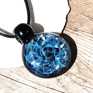 A round memorial glass pendant with a swirling blue with ashes design, resembling a nebula. The pendant is attached to a black cord. The pendant's top is black and rounded. The pendant is resting on a wooden surface.