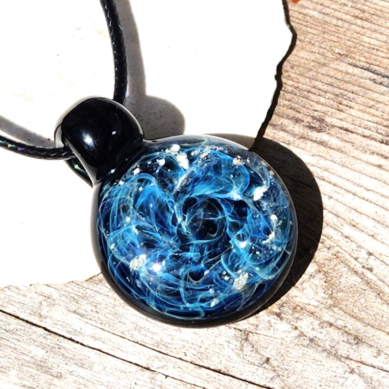 A round memorial glass pendant with a swirling blue with ashes design, resembling a nebula. The pendant is attached to a black cord. The pendant's top is black and rounded. The pendant is resting on a wooden surface.