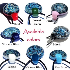 A collection of handmade glass cremation pendants in various colors, including red, forest green, pink, stormy blue, black, white, ocean blue, and sky blue. Each memorial pendant features a unique swirling blue with ashes design and is strung on a black cord. The text "Available colors" is also visible.