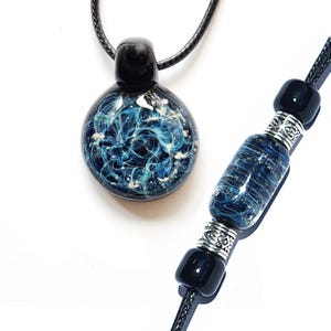May include: A round glass pendant with a swirling blue and white nebula pattern on a black cord, accompanied by a matching bracelet featuring a cylindrical blue glass bead, silver-toned metal spacers, and small black beads on a black cord.