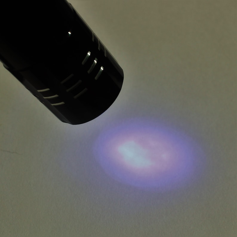 May include: A black flashlight with a grid pattern on the end is shining a bright purple light onto a white surface.