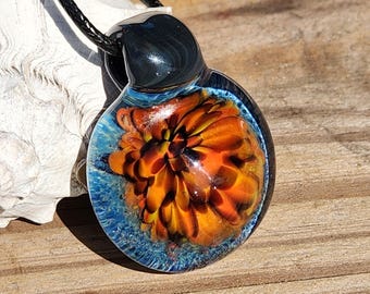 May include: A round glass pendant featuring an orange and yellow floral pattern encased in clear glass with a blue textured base. The pendant has a black glass bail and is suspended from a black cord, creating a circular jewelry piece.