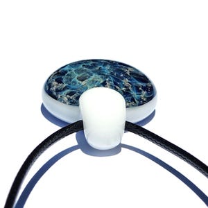 A handcrafted urn pendant necklace featuring a round, blue with mixed in ashes glass pendant with a white bail. The pendant is suspended from a black cord. The glass pendant has a unique, swirling design.