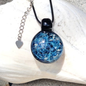 A round glass memorial pendant necklace with a black loop and a blue with ashes swirling design. The pendant hangs from a black cord with a stainless steel silver-toned extension chain and heart-shaped charm. The necklace is displayed on a white surface.