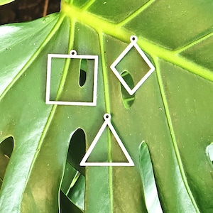 Puede incluir: Tres pendientes geométricos plateados: un cuadrado, un rombo y un triángulo. Cada pendiente tiene un pequeño lazo para colgar. Los pendientes están sobre una gran hoja verde.