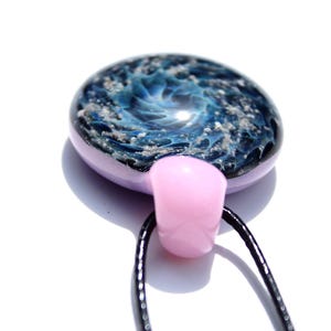 A handcrafted cremation glass pendant with a swirling blue and white design, resembling a galaxy. The pendant has a pink border and a pink bail, suspended from a black cord. The overall design is artistic and unique.