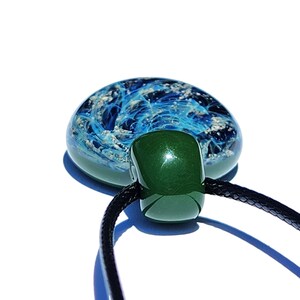 A handcrafted cremation pendant featuring a blue pure silver with infused ashes swirling design encased in clear glass. The pendant is connected to a black cord necklace. The design resembles ocean waves.