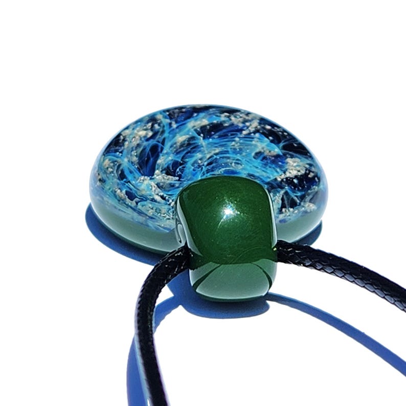 A handcrafted cremation pendant featuring a blue pure silver with infused ashes swirling design encased in clear glass. The pendant is connected to a black cord necklace. The design resembles ocean waves.