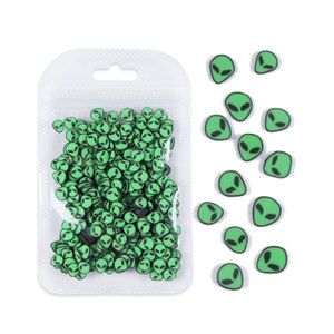Puede incluir: Una bolsa de plástico transparente llena de espolvorear verde en forma de cara de extraterrestre. Se muestra un montón separado de espolvorear verde en forma de cara de extraterrestre sobre un fondo blanco.