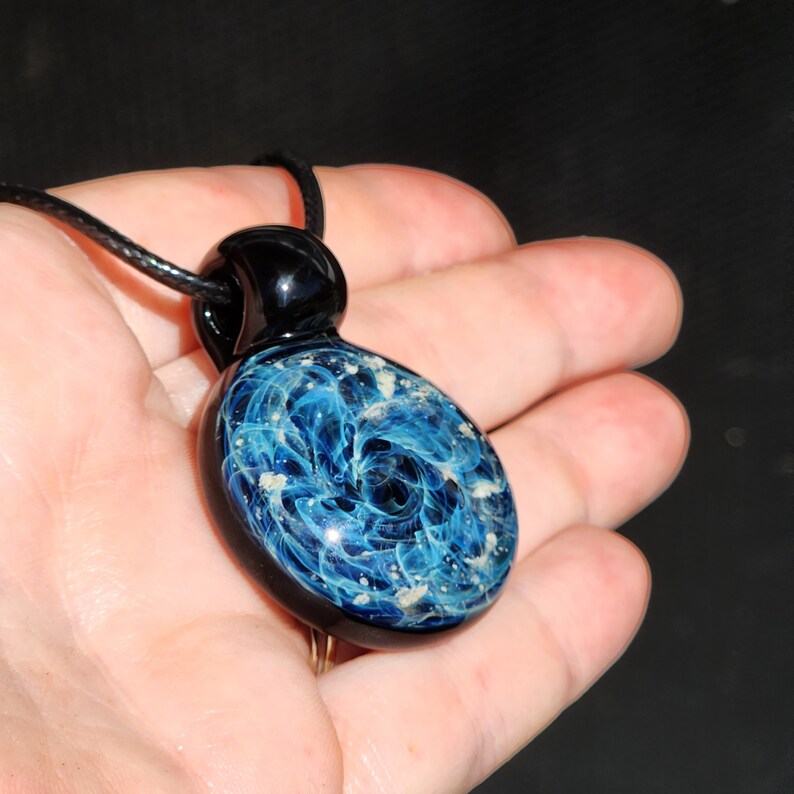 Cremation jewelry glass pendant with a swirling blue and infused ashes design, suspended from a black cord. The pendant has a black top and a glossy finish. The design resembles a galaxy or nebula.