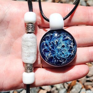 May include: A round glass pendant featuring a deep blue and swirling light blue nebula-like pattern with silver flecks, suspended on a black cord. Beside it, a black cord features two white beads and a textured white bead with silver spacers.
