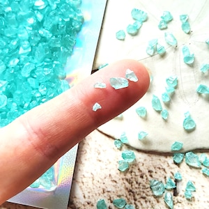 May include: A close-up of turquoise blue glass shards, some of which are being held by a finger. The shards are small and irregular in shape.