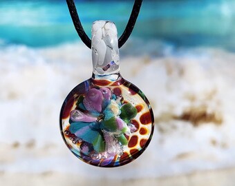 May include: A round glass pendant on a black cord featuring an internal floral-like design with purple, blue, and green petals surrounded by a brown and orange honeycomb pattern, topped with a clear glass bail containing white flecks.