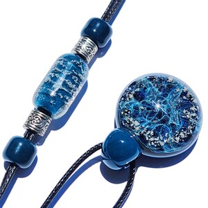 May include: A necklace featuring a round glass pendant with a swirling blue and silver nebula-like pattern, paired with a black cord. The cord is accented with smooth dark blue beads and ornate silver-toned metal spacers, creating a cohesive design.
