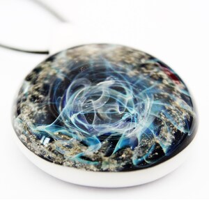 A round memorial pendant with a white border, featuring a swirling blue with pet's ashes design within a clear, domed surface. The design resembles galaxy or a nebula against a dark background.