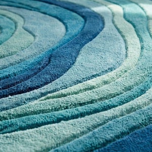 Puede incluir: Primer plano de una alfombra redonda con un patrón de ondas en tonos azules y turquesas. La alfombra tiene una superficie suave y texturizada, con los colores mezclándose para crear un diseño fluido. El diseño de la alfombra recuerda a las olas del océano.