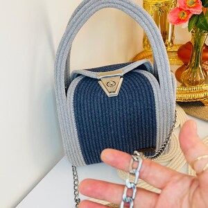 May include: A structured handbag crafted from coiled rope in light gray and dark blue. The bag features a top handle, a silver-toned metal turn-lock closure on the front flap, and a detachable silver-toned metal chain shoulder strap.