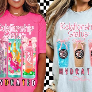 Könnte beinhalten: Zwei T-Shirts, eines in Pink und eines in Hellblau, jeweils mit dem Text "Relationship Status:" in rosa Schrift. Das pinkfarbene Shirt zeigt drei Alani Nu Energy-Drink-Dosen, und das blaue Shirt hat drei Seven Brew Kaffeegetränke. Das Wort "HYDRATED" steht darunter.