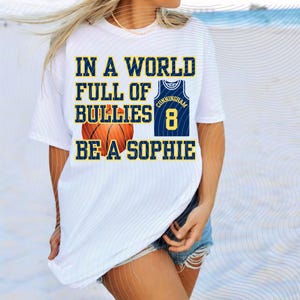 Puede incluir: Camiseta blanca con el texto "IN A WORLD FULL OF BULLIES BE A SOPHIE". El diseño incluye una pelota de baloncesto y una camiseta azul marino con el nombre "Cunningham" y el número "8".